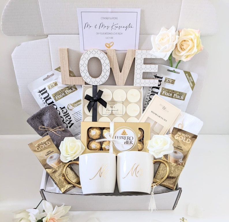 May include: A gift box filled with various items for a wedding celebration. The box includes a wooden LOVE sign, candles, chocolates, coffee, and a towel. The box is decorated with white flowers and a card that reads "Congratulations Mr & Mrs Kensington".