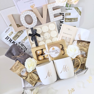 May include: A white gift box filled with wedding-themed gifts, including two white mugs with gold lettering that say "Mr." and "Mrs.", a box of chocolates, a white towel, candles, and a wooden sign that says "LOVE".
