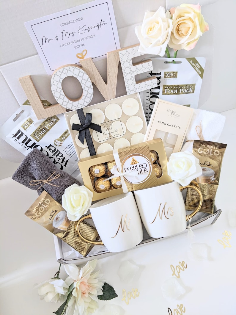 May include: A white gift box filled with wedding-themed gifts, including two white mugs with gold lettering that say "Mr." and "Mrs.", a box of chocolates, a white towel, candles, and a wooden sign that says "LOVE".