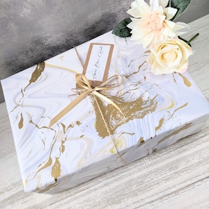 May include: A white gift box with a gold marble pattern, wrapped with a gold ribbon and a brown tag that says "Happy Birthday". The box is sitting on a white wooden surface with a white and peach flower on top.