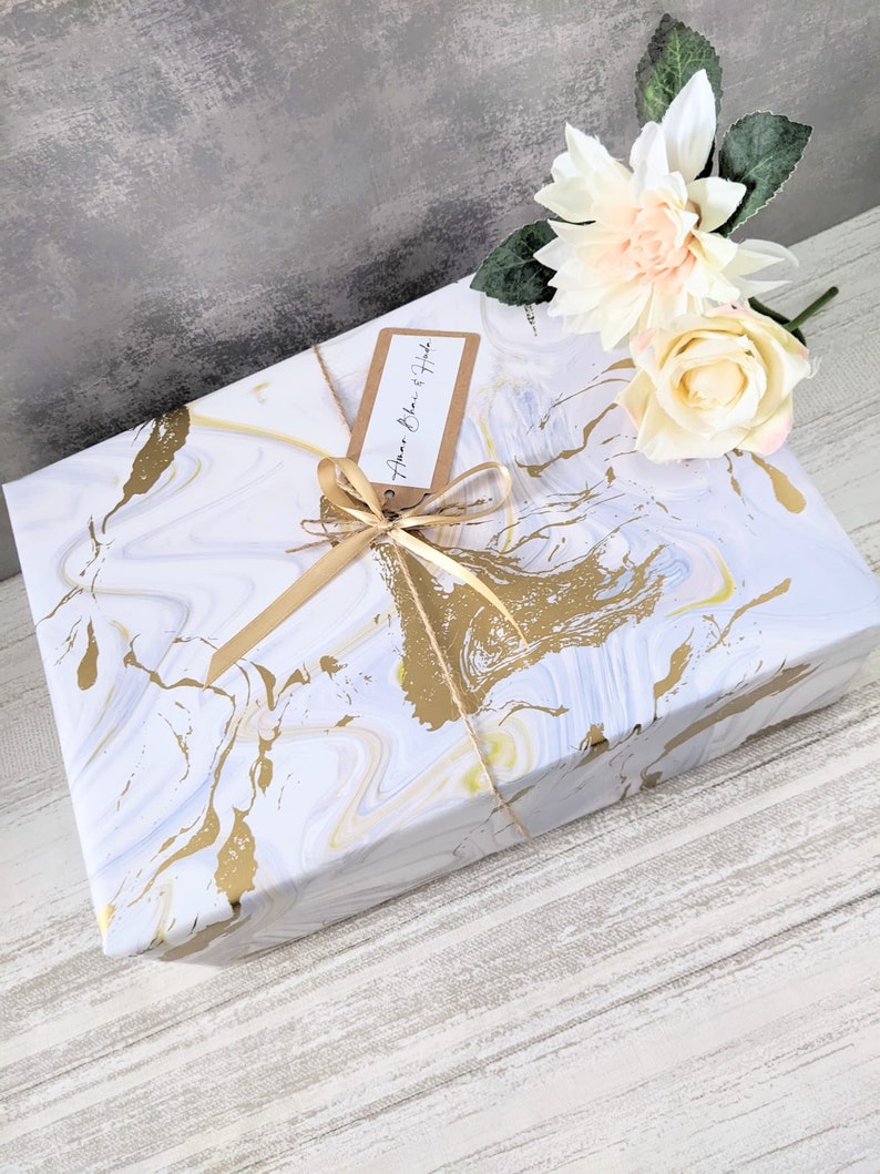 May include: A white gift box with a gold marble pattern, wrapped with a gold ribbon and a brown tag that says "Happy Birthday". The box is sitting on a white wooden surface with a white and peach flower on top.