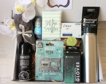 Puede incluir: Una caja de regalo llena de diversos artículos, incluyendo una toalla gris, una botella de spray corporal, una pastilla de jabón, una pequeña botella de desinfectante de manos, un paquete de pañuelos, una tarjeta de crédito multiusos, un par de guantes táctiles, una mascarilla facial de peeling para puntos negros y una botella de agua de acero inoxidable. La caja está decorada con virutas de papel marrón y una tarjeta blanca con el texto "DREAM Believe ACHIEVE".