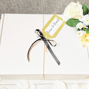 May include: A white gift box with a gold glitter tag that says "Laura & Daniel Congratulations". The box is tied with a black ribbon and twine.