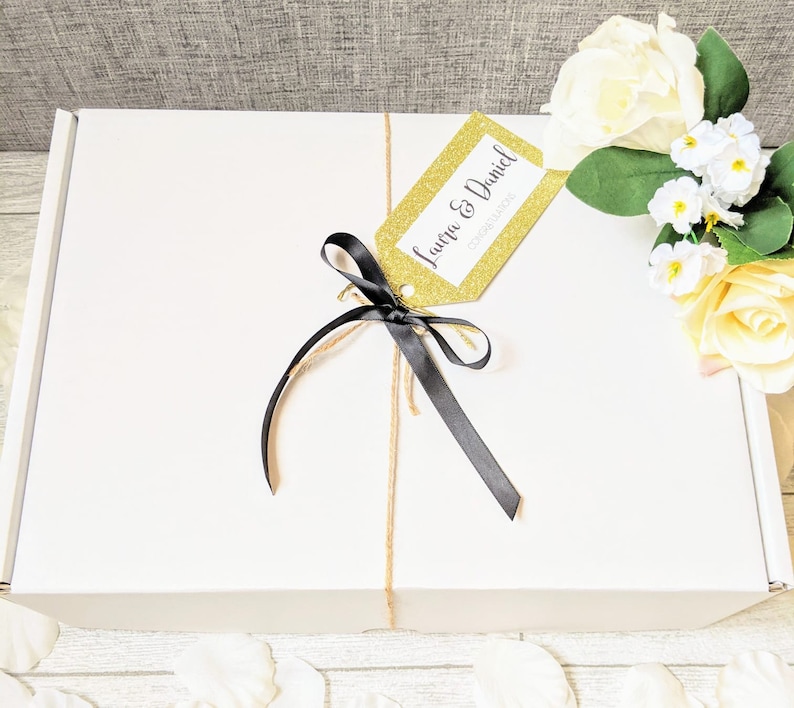 May include: A white gift box with a gold glitter tag that says "Laura & Daniel Congratulations". The box is tied with a black ribbon and twine.