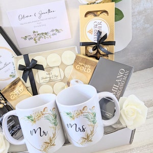 May include: A gift box filled with wedding-themed items, including two white mugs with gold accents and the words "Mr" and "Mrs" on them, a box of chocolates, candles, and a card that reads "Olivia & Jonathan, May the years ahead be filled with lasting joy, From Emma & Doug".