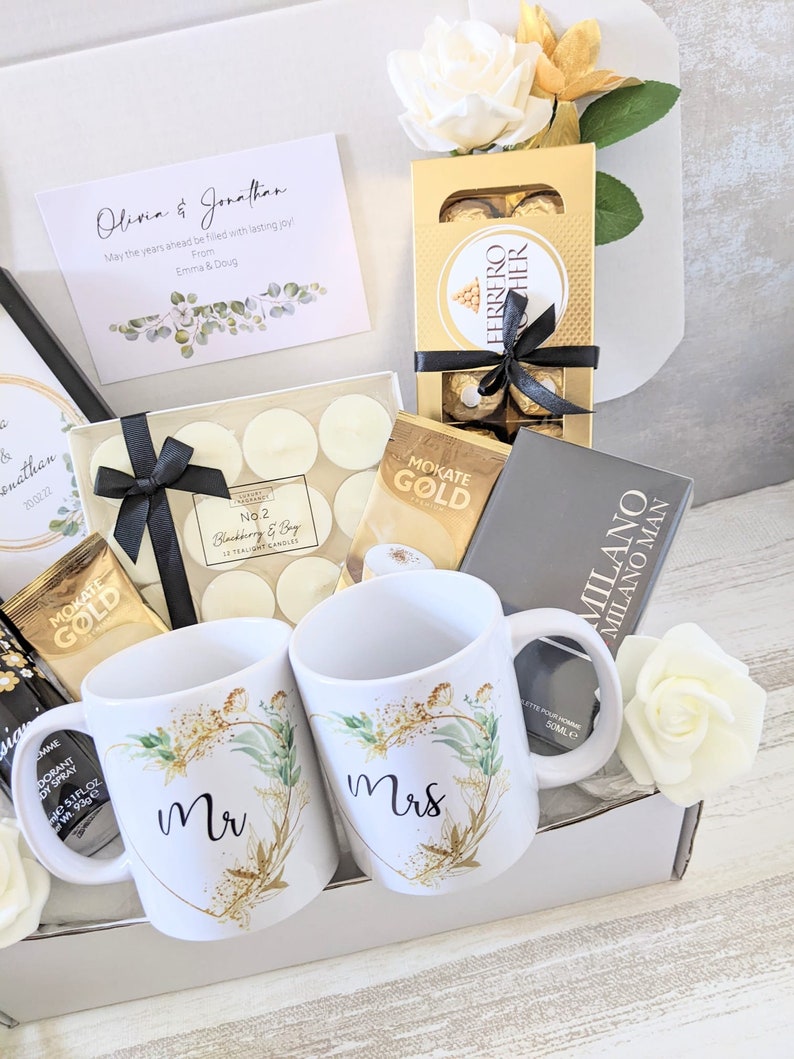 May include: A gift box filled with wedding-themed items, including two white mugs with gold accents and the words "Mr" and "Mrs" on them, a box of chocolates, candles, and a card that reads "Olivia & Jonathan, May the years ahead be filled with lasting joy, From Emma & Doug".