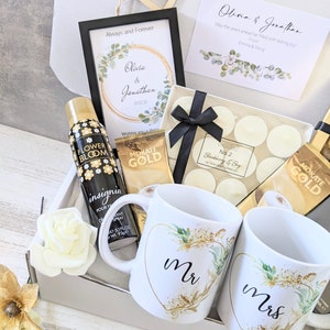 May include: A gift box filled with wedding-themed items, including two white mugs with gold accents and the words "Mr." and "Mrs." on them, a bottle of body spray, a box of tea, and a box of candles.