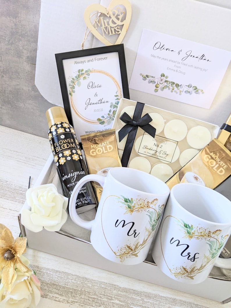 May include: A gift box filled with wedding-themed items, including two white mugs with gold accents and the words "Mr." and "Mrs." on them, a bottle of body spray, a box of tea, and a box of candles.