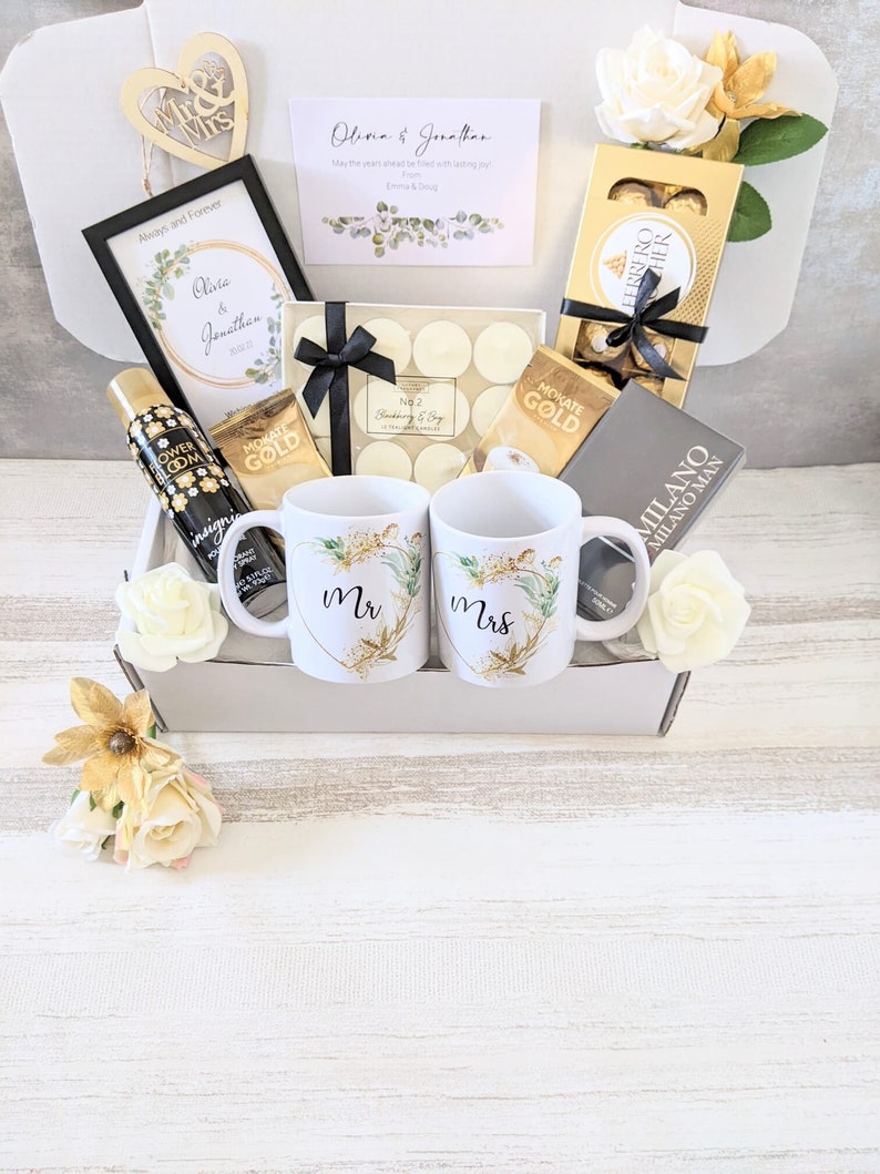 May include: A white gift box filled with wedding-themed gifts, including two mugs with "Mr." and "Mrs." on them, a framed print with the words "Olivia & Jonathan", a box of chocolates, and a bottle of body spray.