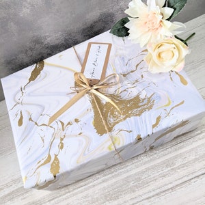 May include: A white gift box with a gold marble pattern, wrapped with a gold ribbon and a brown tag that says "Happy Birthday". The box is sitting on a white wooden surface with a white and peach flower on top.