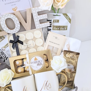 May include: A gift basket filled with wedding-themed items, including two white mugs with gold lettering that say "Mr." and "Mrs.", a box of chocolates, a foot mask, candles, and a bag of coffee. The basket is decorated with white roses and the word "LOVE" in wooden letters.