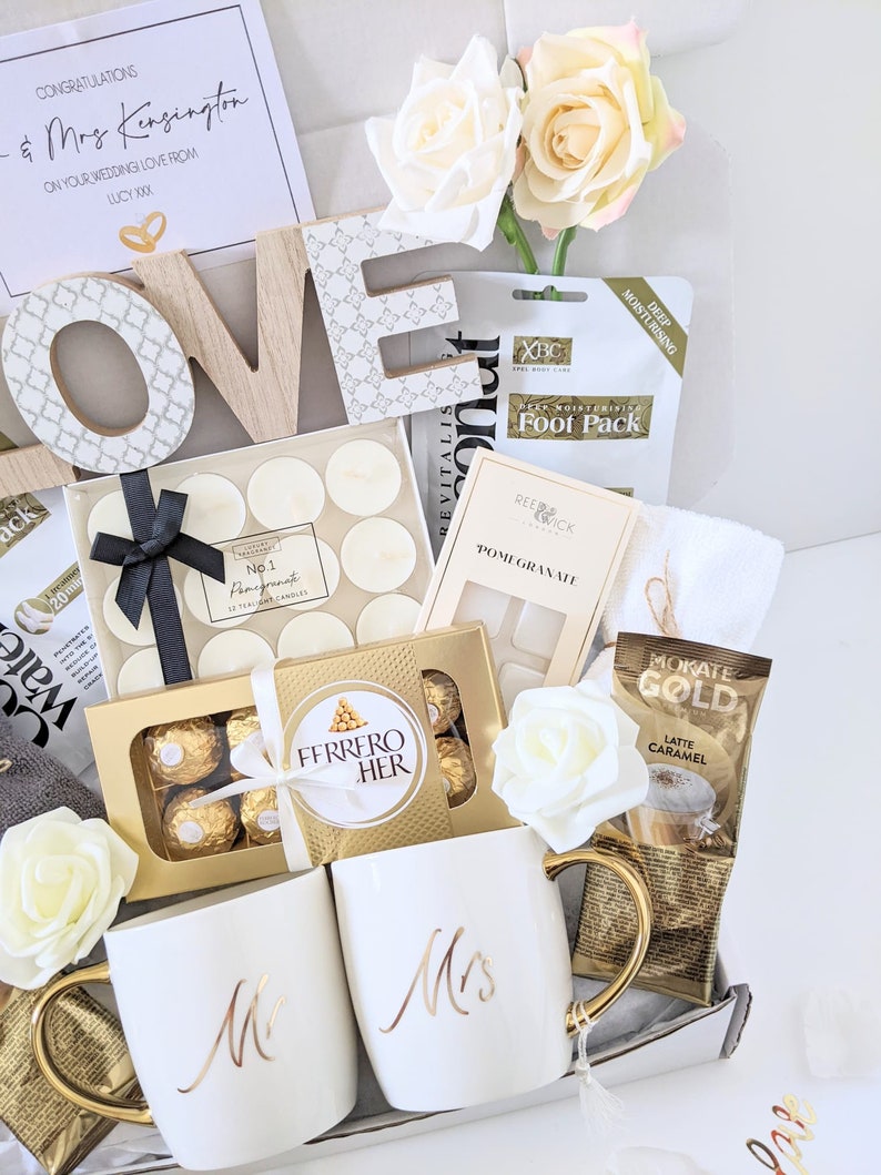 May include: A gift basket filled with wedding-themed items, including two white mugs with gold lettering that say "Mr." and "Mrs.", a box of chocolates, a foot mask, candles, and a bag of coffee. The basket is decorated with white roses and the word "LOVE" in wooden letters.