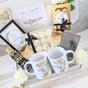 May include: A gift basket filled with wedding-themed items, including two white mugs with gold accents and the words "Mr." and "Mrs." on them, a box of chocolates, a candle, a bottle of body spray, and a card with the words "Olivia & Jonathan" on it.