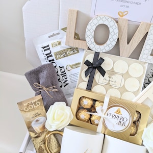 May include: A gift box filled with wedding gifts for a couple. The box contains two white mugs with gold lettering that says "Mr" and "Mrs", a box of chocolates, a box of tea lights, a foot pack, a packet of coffee, and a wooden sign that says "LOVE".