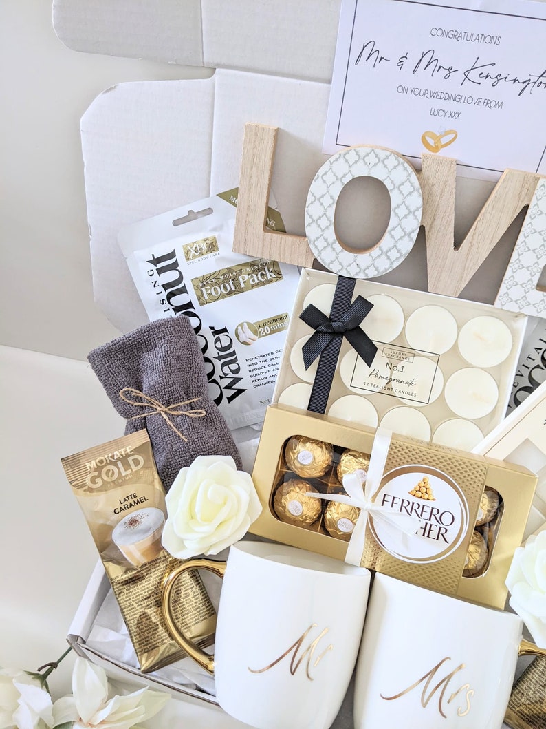 May include: A gift box filled with wedding gifts for a couple. The box contains two white mugs with gold lettering that says "Mr" and "Mrs", a box of chocolates, a box of tea lights, a foot pack, a packet of coffee, and a wooden sign that says "LOVE".
