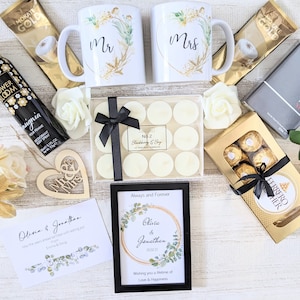 May include: A wedding gift basket with a variety of items, including two white mugs with gold accents and the words "Mr." and "Mrs." on them, a box of white candles, a box of chocolates, a framed print with a floral design and the text "Olivia & Jonathan", a card with the text "Olivia & Jonathan", a wooden heart with the words "Mr. & Mrs." on it, and a bottle of body spray.