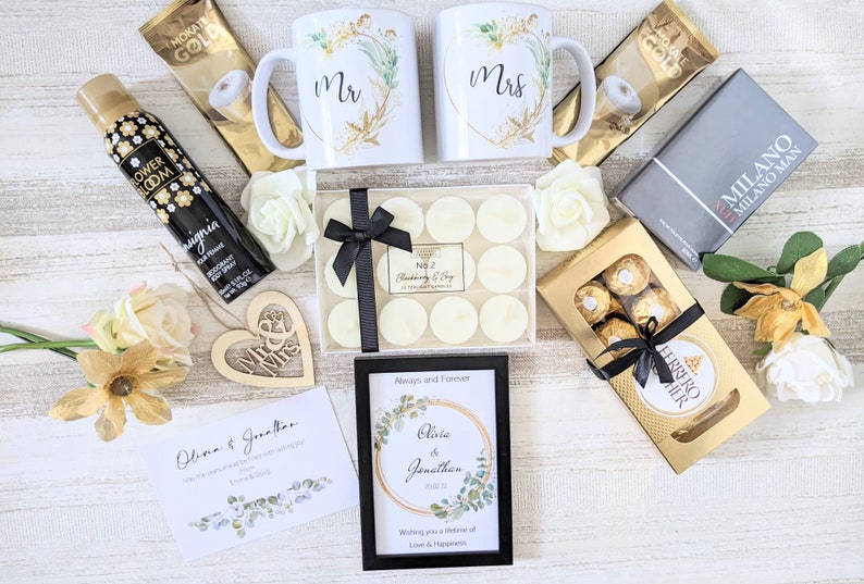 May include: A wedding gift basket with a variety of items, including two white mugs with gold accents and the words "Mr." and "Mrs." on them, a box of white candles, a box of chocolates, a framed print with a floral design and the text "Olivia & Jonathan", a card with the text "Olivia & Jonathan", a wooden heart with the words "Mr. & Mrs." on it, and a bottle of body spray.