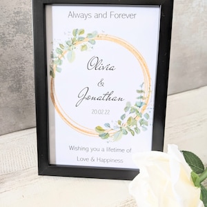 May include: A black framed print with a gold and green wreath design. The text reads "Always and Forever Olivia & Jonathan 20.02.22 Wishing you a lifetime of Love & Happiness".