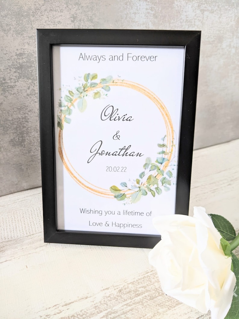 May include: A black framed print with a gold and green wreath design. The text reads "Always and Forever Olivia & Jonathan 20.02.22 Wishing you a lifetime of Love & Happiness".