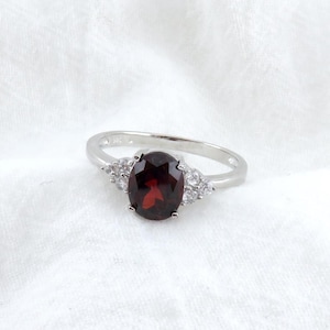 May include: A silver ring with a red oval gemstone surrounded by smaller clear gemstones.