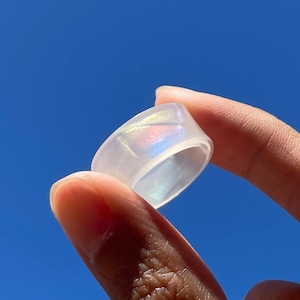May include: A white iridescent ring with a smooth, polished finish. The ring is held up against a bright blue sky.