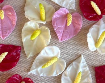 May include: An overhead shot displays numerous artificial anthurium flowers in various colors. The flowers are white, pink, and deep red, each with a yellow spadix. The arrangement is scattered across a neutral-toned surface.