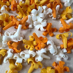 May include: A collection of small, plastic flower-shaped components in shades of white, yellow, and orange. The components have a detailed, three-dimensional design, with curved petals and a central stem. The pieces are scattered on a neutral-colored surface.