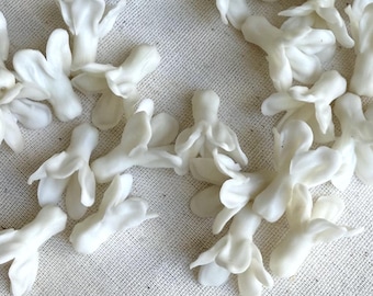 May include: A collection of small, white, flower-shaped embellishments scattered on a light-colored surface. The delicate, three-dimensional floral components have a smooth, glossy finish and are clustered together, suggesting a craft or decorative theme.