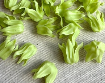 May include: A collection of small, light green flower buds scattered on a neutral-toned fabric. The buds have a delicate, layered petal design and a slightly glossy finish, suggesting a craft or decorative item.