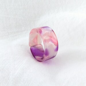 May include: A pink and purple marbled resin ring. The ring is wide and has a smooth, polished finish.