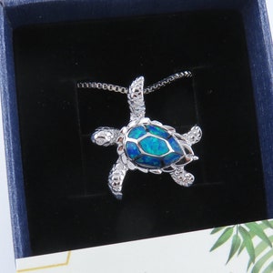 May include: A silver-toned turtle pendant necklace with a blue and teal-colored center stone, presented in a navy blue box. The necklace is on a silver chain. The box contains a card with the text "Seaside Avenue".