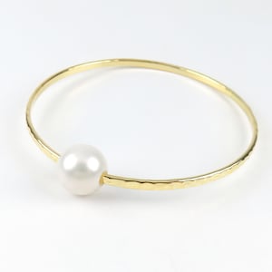 May include: A gold bangle bracelet with a single white pearl accent. The bracelet has a hammered texture.