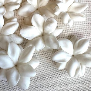 May include: A collection of eight white paper flowers with five petals each. The flowers are arranged in a cluster on a white fabric background.