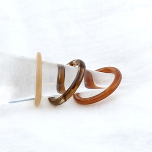 May include: Two rings, one brown and one beige, are displayed on a clear plastic cone. The rings are made of a smooth, glossy material.