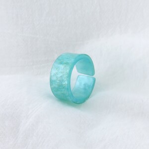 May include: A turquoise blue ring with a sparkly finish. The ring is made of resin and has a simple, modern design.