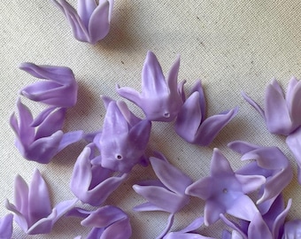 Puede incluir: Una colección de pequeños capullos de flores de color lavanda esparcidos sobre una tela beige claro. Los capullos están detallados con múltiples pétalos, y la disposición general sugiere un tema artesanal o decorativo.