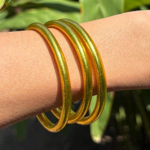 May include: Three gold-colored bangle bracelets stacked on a person's wrist. The bracelets are shiny and have a metallic finish.