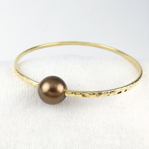 Puede incluir: Un brazalete de oro con textura martillada y una sola perla marrón.