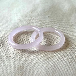 Puede incluir: Dos anillos de jade rosa pálido, uno ligeramente más grande que el otro, están sobre una superficie de tela blanca.