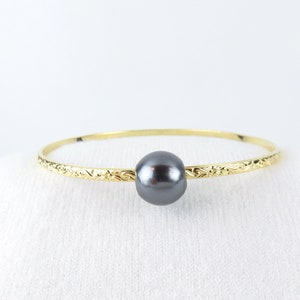 May include: A gold bangle bracelet with a single black pearl centered on the band. The band has a delicate, engraved design.