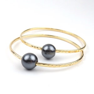 May include: Two gold-colored bangles with a hammered texture, each adorned with a dark gray pearl. The jewelry is set against a white background, highlighting the contrast between the gold and the pearls. A simple and elegant design.