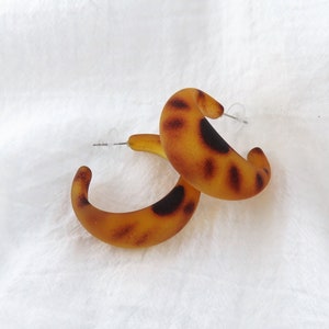 May include: A pair of hoop earrings with a tortoiseshell pattern. The earrings are a warm amber color with dark brown spots. The earrings are made of a translucent material and have a post closure. The earrings are on a white background.