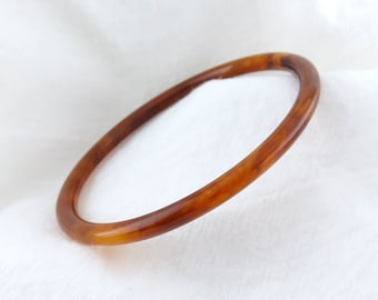 May include: A thin, circular bangle bracelet featuring a translucent, mottled amber-brown color with a smooth, polished finish. The bracelet has a simple, continuous band design with a rounded profile, showcasing variations in tone throughout.