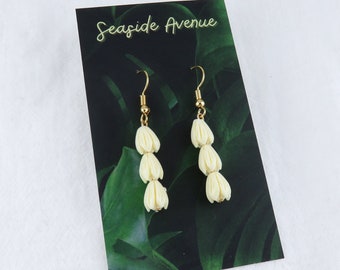 Puede incluir: Un par de pendientes colgantes de flores blancas con ganchos dorados. Los pendientes se muestran en una tarjeta con el texto "Seaside Avenue".
