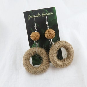 May include: A pair of brown, rope circle earrings with a brown, textured bead on each earring. The earrings are hanging from silver hooks. The earrings are on a white background with a card that says "Seashore Avenue".