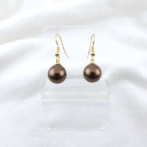May include: A pair of brown pearl earrings with gold-colored hooks. The earrings are displayed on a clear acrylic stand against a white background. The pearls are round and have a lustrous finish.