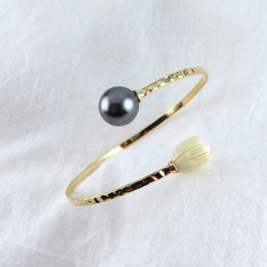 May include: A gold bangle bracelet with a hammered texture. The bracelet has a black pearl on one end and a white flower-shaped charm on the other end.