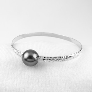 May include: A silver bangle bracelet with a dark gray pearl charm. The bracelet is decorated with a floral design.