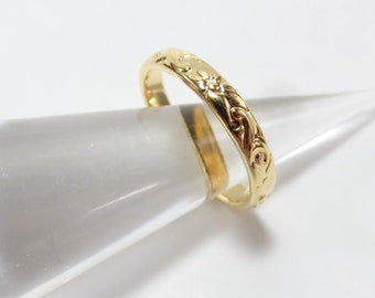 May include: A gold band ring with an intricate floral design. The ring is on a clear display stand.