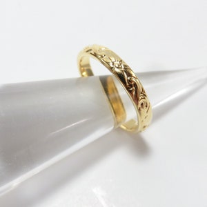 May include: A gold band ring with an intricate floral design. The ring is on a clear display stand.
